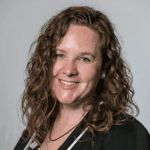  A woman with Shoudler length hair, with a black blazer on who smiles at the camera in front of a grey background. 