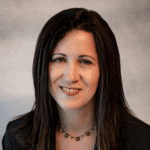  A woman with long brunette hair, burgundy necklace, a black cardigan smiles at the camera in front of a cloudy grey blurred background. 